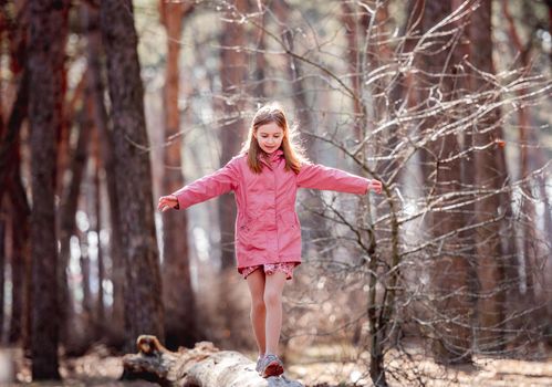 Little beautiful girl wearing pink coat walks on a log in the wood with sunlight in her hair