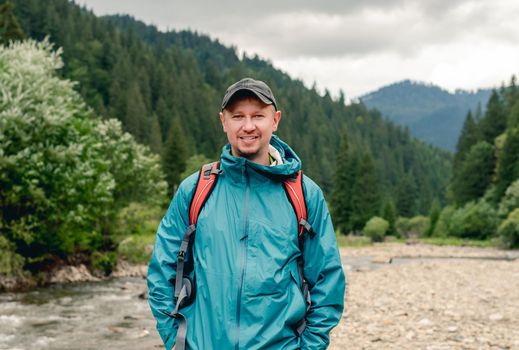 Smiling young man tourist with backpack standing on river mountain background