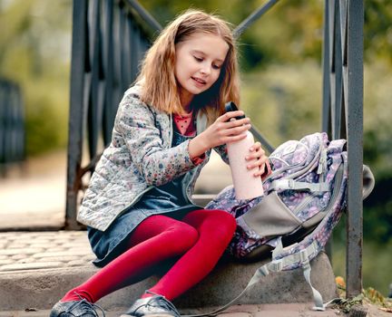Preteen school girl with backpack sitting on ground and holding bottle with water. Sweet pupil female kid resting after education class in autumn park