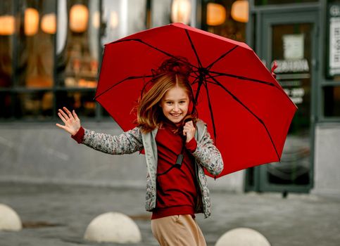 Preteen girl wearing roober boots and holding umbrella jumping in rainy autumn day and smiling. Pretty child in gumboots having fun at wet street