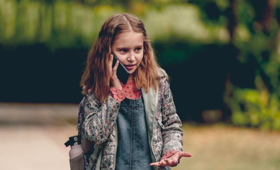 Pretty school girl talking by smartphone outdoors and smiling. Cute female kid communicates using cell phone in autumn park