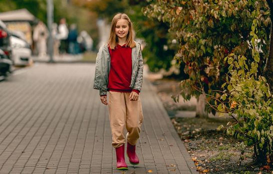 Preteen girl wearing rubber boots walking at autumn at street. Pretty female kid in gumboots outdoors