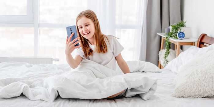 Girl talking by video call using smartphone in the bed and smiling. Female kid with cell phone in the morning time in the room with daylight