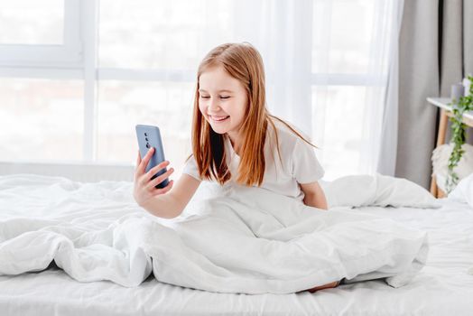 Little girl making selfie using smartphone in the bed and smiling. Female kid with cell phone in the morning time in the room with daylight. Kid and technologies