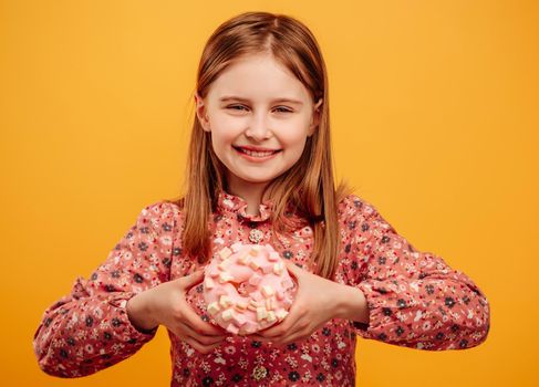 Little girl holding donut and looking at the camera isolated on yellow background with copyspace. Pretty female kid with sweets during studio photosession