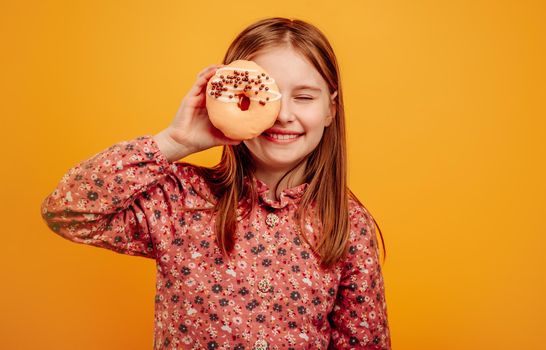 Little girl playing and holding donut as glasses close to eyes isolated on yellow background with copyspace. Pretty female kid with sweets