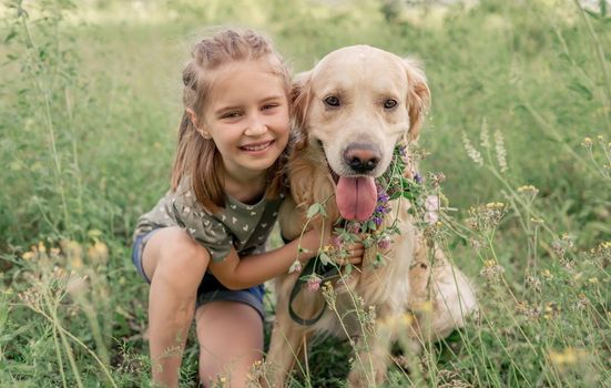 Portrait of beautiful preteen girl hugging golden retriever dog and smiling looking at the camera outdoors. Cute female kid with doggy pet sitting at the nature in summer time in the grass