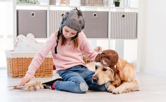 Little girl in hat playing with wooden plain and golden retriever dog in pilot glasses lying next to her