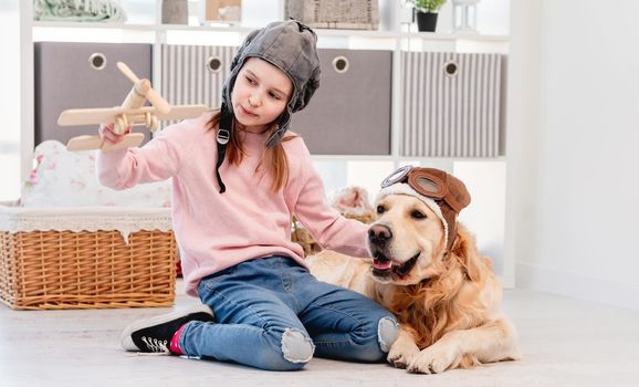 Little beautiful girl and golden retriever dog wearing pilot costumes playing games with wooden plain