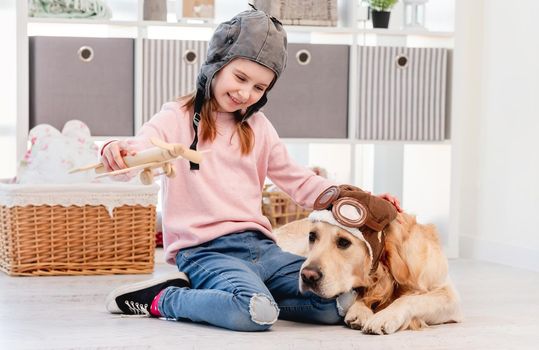 Little girl playing with wooden plain and golden retriever dog wearing pilot glasses lying on the floor