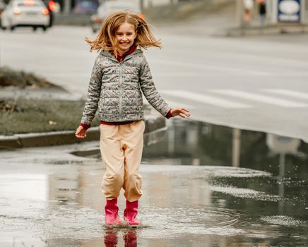 Preteen girl wearing rubber boots jumping through puddles at autumn at street. Pretty female kid in gumboots in rainy day outdoors