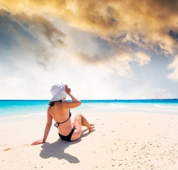 girl in hat and black swimsuit sitting on beautiful beach with blue ocean and sky on the background