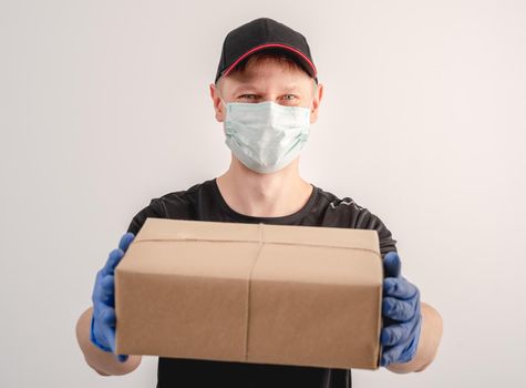 Young courier in protective mask and gloves holding small parcel on white wall background