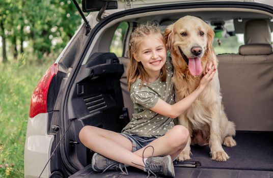 Beautiful little girl sitting with golden retriever dog in the car trunk and smiling looking at the camera. Child kid hugging purebred doggy pet in the vehicle at the nature