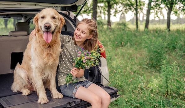 Beautiful little girl sitting with golden retriever dog in the car trunk and holding flower bouquet. Smiling child kid with purebred doggy pet and summer plants in the vehicle at the nature