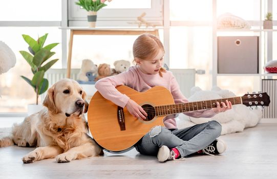 Little girl playing guitar and sitting on the floor with golden retriever dog