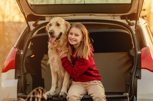 Girl child hugging golden retriever dog sitting in car trunk outdoors. Pretty kid petting doggy pet labrador and smiling in vehicle at nature