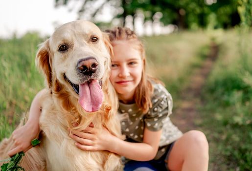 Portrait of beautiful preteen girl with golden retriever dog looking at camera together, hugging and smiling outdoors. Kid with doggy pet in the field in summer time