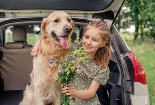 Beautiful little girl sitting with golden retriever dog in the car trunk, hugging it and holding flower bouquet. Child kid with purebred doggy pet and summer plants in the vehicle at the nature