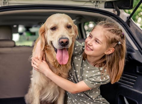 Beautiful little girl sitting with golden retriever dog in the car trunk and smiling looking at the camera. Child kid hugging purebred doggy pet in the vehicle at the nature
