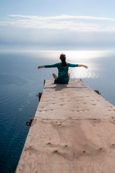 A girl sits on a wooden springboard for jumping with a rope. In a dark green dress with her hands in the air