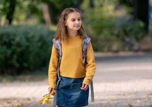 Preteen school girl with backpack holding yellow leaves in her hand and walking in autumn park. Pretty pupil female kid portrait outdoors