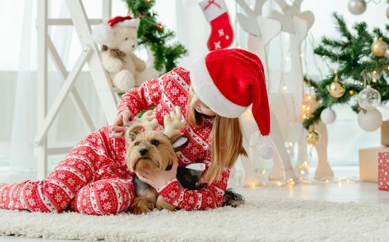 Child girl with dog at Christmas time at home with holiday decoration. Kid wearing red costume and pet doggy in room with New Year decoration