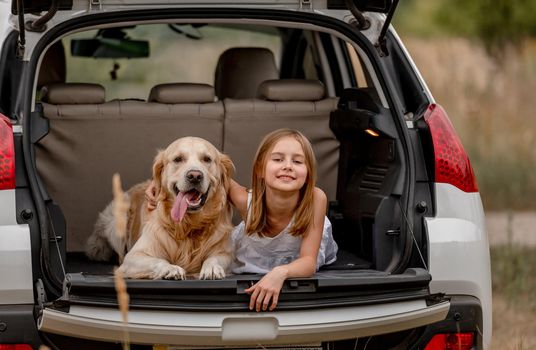 Pretty little girl with golden retriever dog lying in car trunk together and looking at camera. Cute child kid resting with doggy pet in vehicle