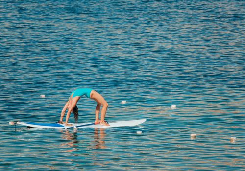 Young woman in blue swimsuite doing yoga on sup board with paddle. Balanced pose - concept of harmony with the nature, free and healthy living, freelance, remote business