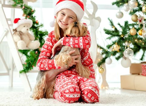 Child girl hugging dog near Christmas tree and happy smiling. Kid wearing Santa hat celebrating New Year with doggy pet terrier