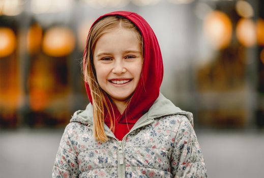 Preteen girl wearing hood and smiling at the street at autumn. Pretty female kid portrait outdoors in fall season