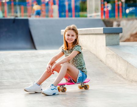 Beautiful girl sitting on skateboard outdoors and smiling in the park. Female skater child close to city riding ramp