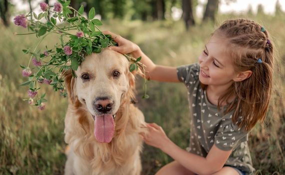 Portrait of beautiful preteen girl petting golden retriever dog and looking at him outdoors. Kid with doggy pet in the field in summer time