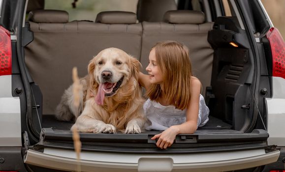 Pretty little girl hugging golden retriever dog and lying in car trunk together. Cute child kid resting with doggy pet in vehicle