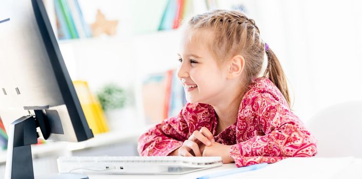 Happy little girl looking at computer screen sitting at desk in classroom