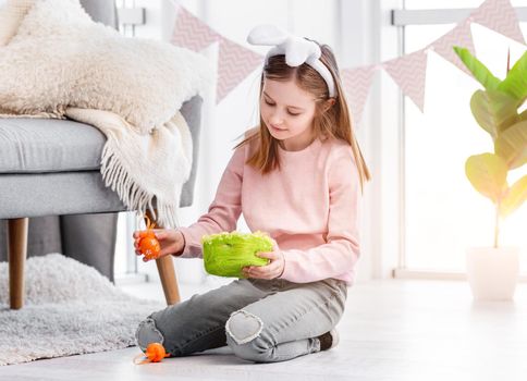 Little girl sitting on the floor at home and holding green basket and painted egg in her hands at Easter day