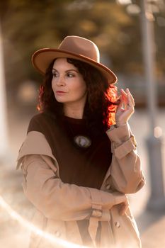 Outdoor fashion portrait of young elegant fashionable brunette woman, model in stylish hat, choker and light raincoat posing at sunset in European city