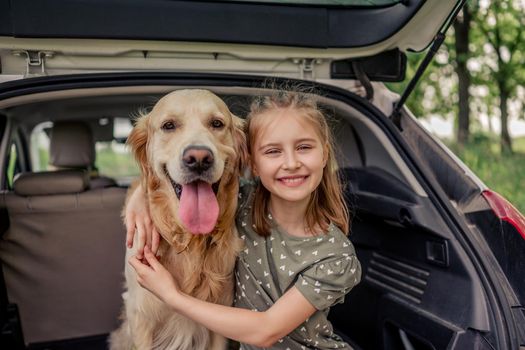 Beautiful little girl sitting with golden retriever dog in the car trunk and smiling looking at the camera. Child kid hugging purebred doggy pet in the vehicle at the nature