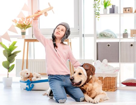 Little girl wearing pilot hat sitting on the floor with golden retriever dog in pilot glasses and play with wooden plane