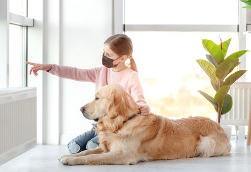 Little girl in black mask sitting with golden retriever dog on the floor and showing him something on the street