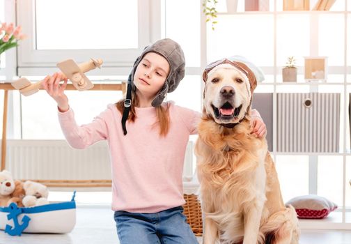 Portrait of little girl and golden retriever dog wearing special pilot glasses and hat during role game