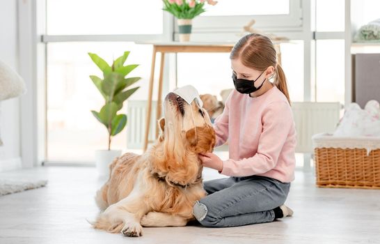 Little girl in black mask sitting on the floor in sunny room and petting golden retriever dog