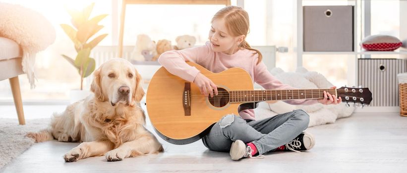 Little girl with guitar sitting on the floor in sunny room with golden retriever dog