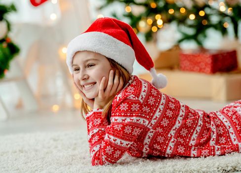 Pretty child girl lying on floor at Christmas home wearing traditional Santaa hat. Happy smiling kid in New Year time