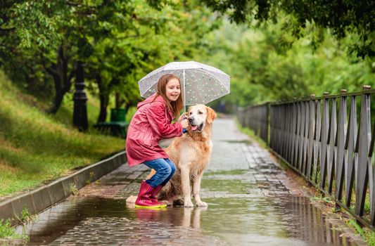 Little girl sharing umbrella with golden retriever standing in puddle on pavement