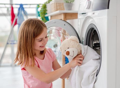 Happy little girl hugging teddy bear in towel after washing on washing machine background