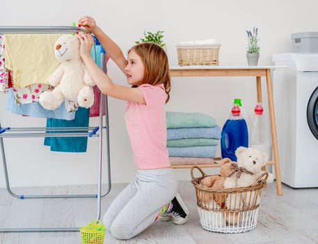 Little girl hanging teddy bear on dryer with pin in light laundry room