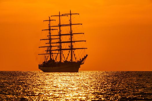 A red burning sunset with the silhouette of sailing ship. Sailors set sails on masts of ship. White sailing vessel floating in the sea. Copy space