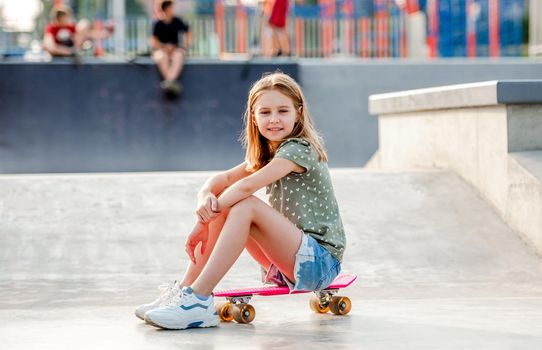 Preteen girl sitting on skateboard outdoors and looking at camera Female skater child close to city riding ramp at summer