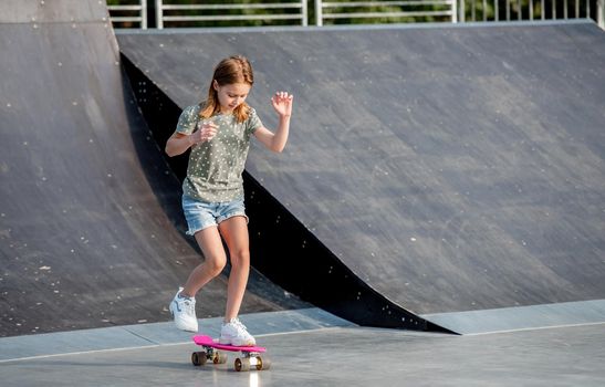 Preteen girl riding on skateboard at city park ramp. Beautiful female child with board outdoors at summer
